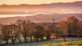 Blick über Kirche mit Zwiebelturm auf See und Berge in Dunst, Herbststimmung