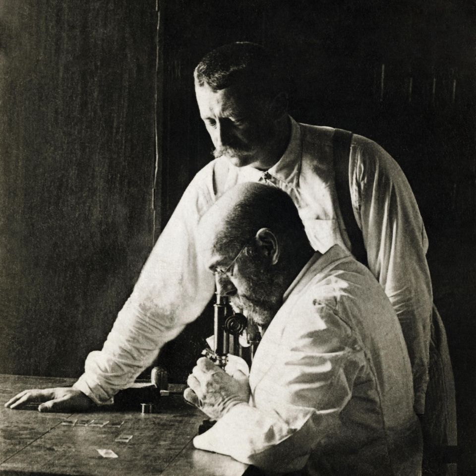 Robert Koch schaut in ein Mikroskop, neben ihm ein Mitarbeiter