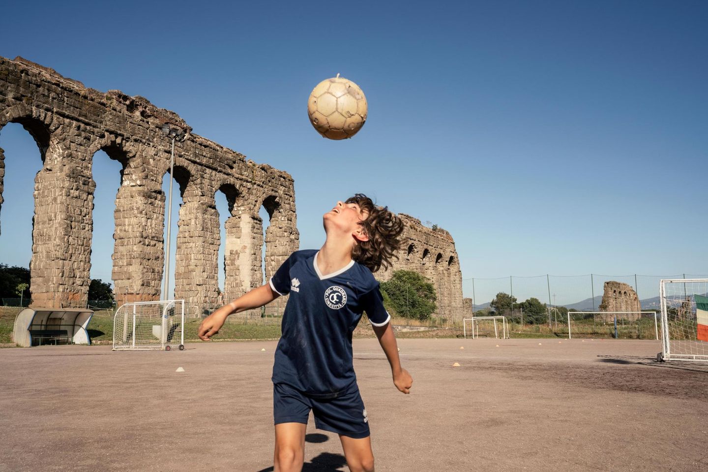 Junge spielt vor den Ruinen eines Aquädukts Fußball