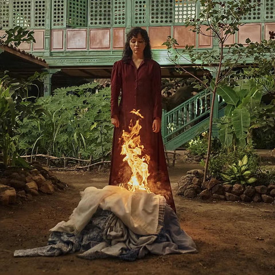 Eine Frau steht in einem Innenhof, vor ihr ein brennender Kleiderhaufen