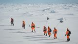 Eine weite Eislandschaft mit Horizont. Sieben Forscher in orangefarbenen Overalls laufen hintereinander.