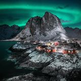Ein Fischerdorf auf den Felsen, nah am Wasser gelegen. Es ist durch grüne Nordlichter am Himmel erleuchtet.