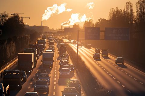 Stau auf der Autobahn bei Sonnenaufgang. Im Hintergrund Emissionen aus Industrieschloten.