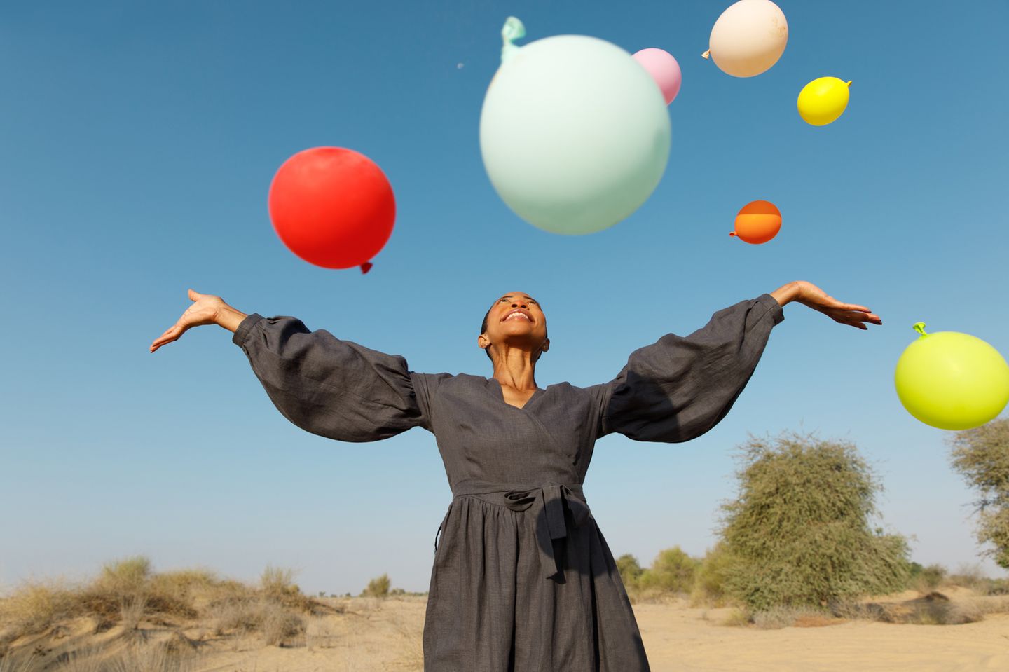 Eine lächelnde Frau im Kleid mit über ihr schwebenden bunten Luftballons vor blauem Himmel.