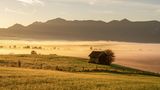 Eine Hütte in einer nebeligen Landschaft im Morgenlicht, im Hintergrund ein Bergpanorama