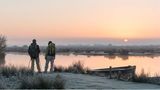 Zwei Wanderer*innen mit Sack und Pack, stehen bei Sonnenaufgang am Wasser des Moores