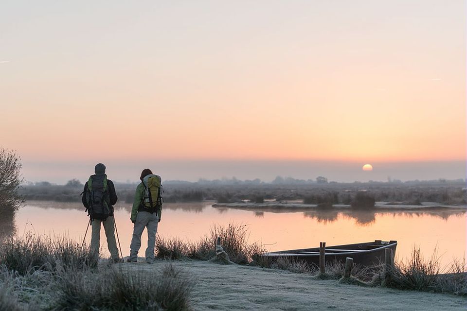 Zwei Wanderer*innen mit Sack und Pack, stehen bei Sonnenaufgang am Wasser des Moores 1.4993