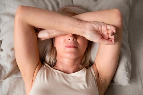 Reife Frau im Bett, die sich mit den Armen die Augen verdunkelt