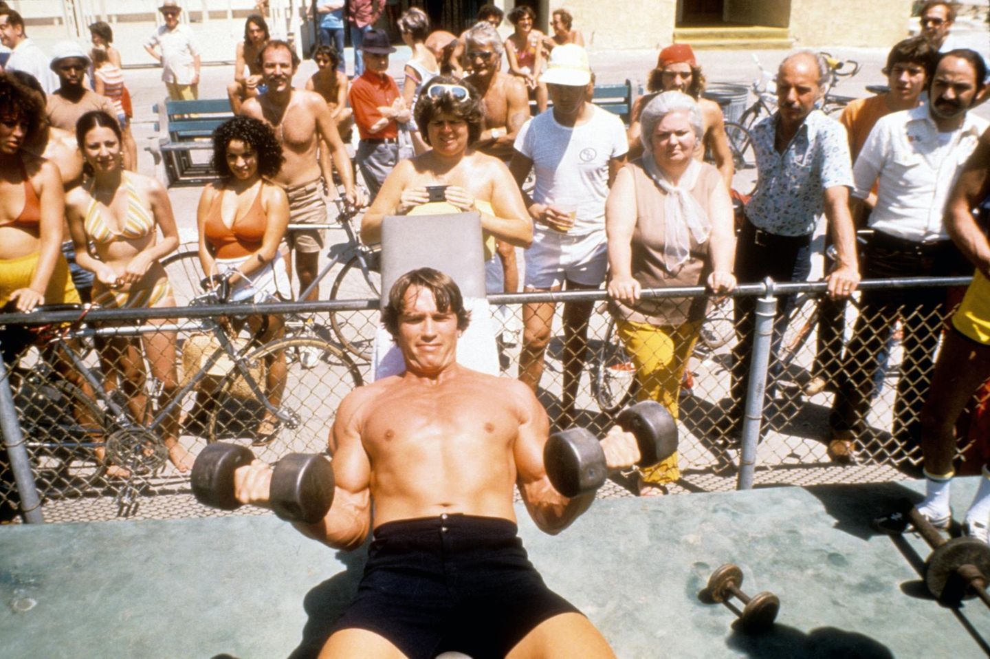 Ein Gigant der Szene Arnold Schwarzenegger stemmt auf dem Rücken liegend zwei Hanteln, hinter einem Zaun schauen Menschen zu