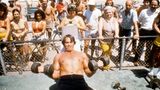 Arnold Schwarzenegger stemmt auf dem Rücken liegend zwei Hanteln, hinter einem Zaun schauen Menschen zu