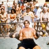 Arnold Schwarzenegger stemmt auf dem Rücken liegend zwei Hanteln, hinter einem Zaun schauen Menschen zu
