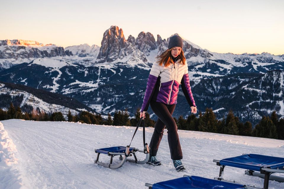 Frau zieht Schlitten über Schnee, im Hintergrund felsige Bergspitzen 1.5026