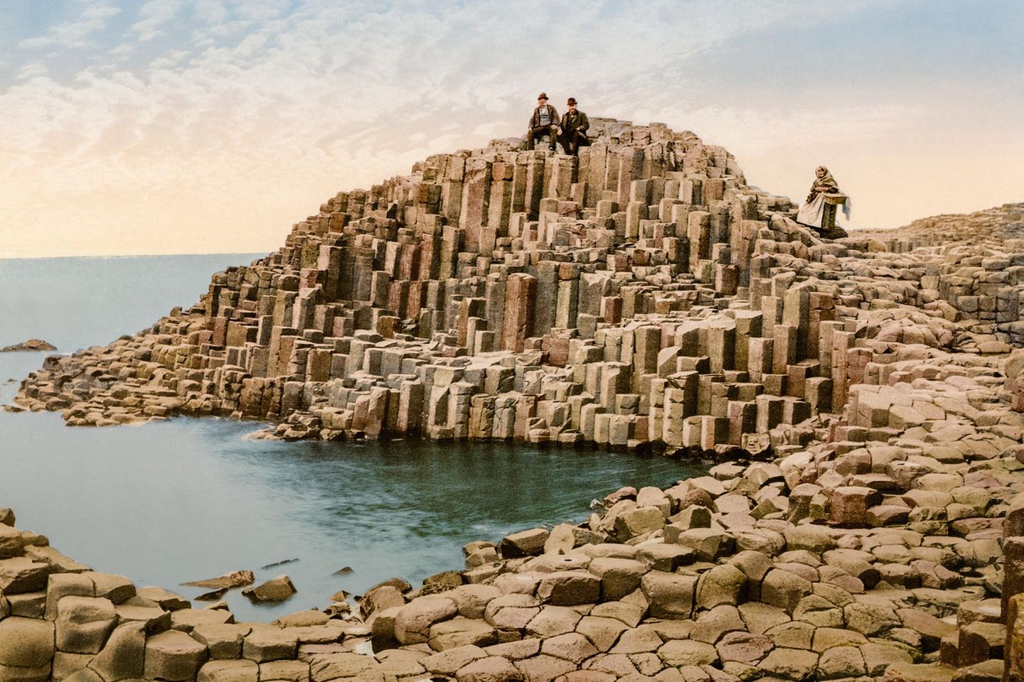 Touristen und eine Verkäuferin auf dem Giant's Causeway, Irland Heute handelt es sich beim Giant's Causeway ("Straße der Riesen") an der nordirischen Küste um eine UNESCO-Weltkulturerbestätte. Die gleichmäßig geformten Steinformationen oder Basaltsäulen sind etwa 60 Millionen Jahre alt und übersähen die Landschaft bis in den Atlantik hinein – als wären sie Trittsteine für Riesen. Bereits um 1900 handelte es sich um ein attraktives Ausflugsziel für Touristen.