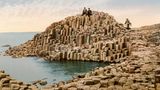 Touristen und eine Verkäuferin auf dem Giant's Causeway, Irland Heute handelt es sich beim Giant's Causeway ("Straße der Riesen") an der nordirischen Küste um eine UNESCO-Weltkulturerbestätte. Die gleichmäßig geformten Steinformationen oder Basaltsäulen sind etwa 60 Millionen Jahre alt und übersähen die Landschaft bis in den Atlantik hinein – als wären sie Trittsteine für Riesen. Bereits um 1900 handelte es sich um ein attraktives Ausflugsziel für Touristen.