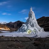 Eine Eisstupa ragt Dutzende Meter in die Höhe. Am Erdboden ist ein kleines Loch eingelassen, darin sitzen Menschen.