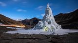 Eine Eisstupa ragt Dutzende Meter in die Höhe. Am Erdboden ist ein kleines Loch eingelassen, darin sitzen Menschen.