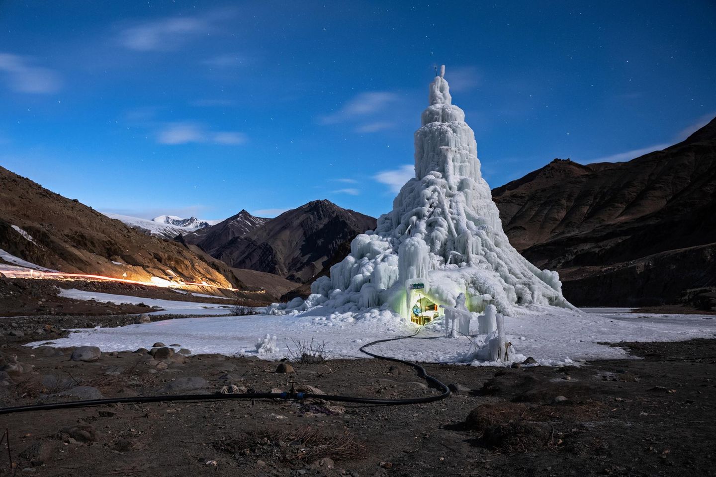 Eine Eisstupa ragt Dutzende Meter in die Höhe. Am Erdboden ist ein kleines Loch eingelassen, darin sitzen Menschen.