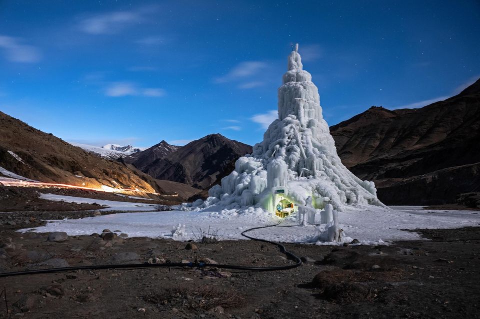 Eine Eisstupa ragt Dutzende Meter in die Höhe. Am Erdboden ist ein kleines Loch eingelassen, darin sitzen Menschen. 1.4993