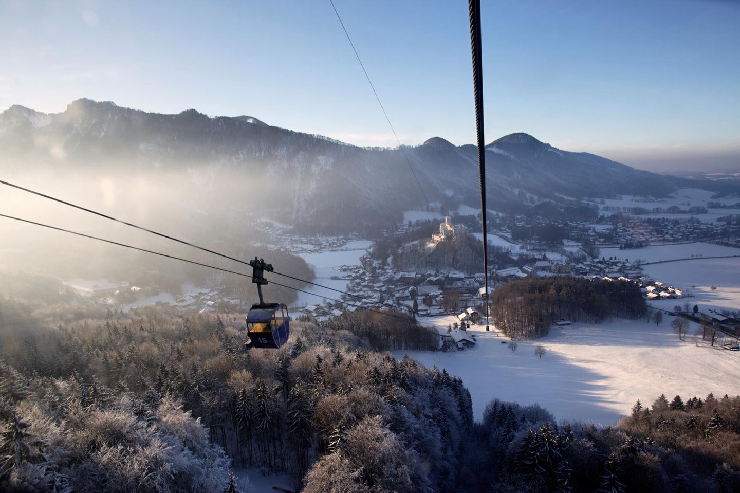 Blick aus einer Gondel der Kampenwandbahn auf verschneite Wälder und Schloss Hohenaschau in der Abensonne