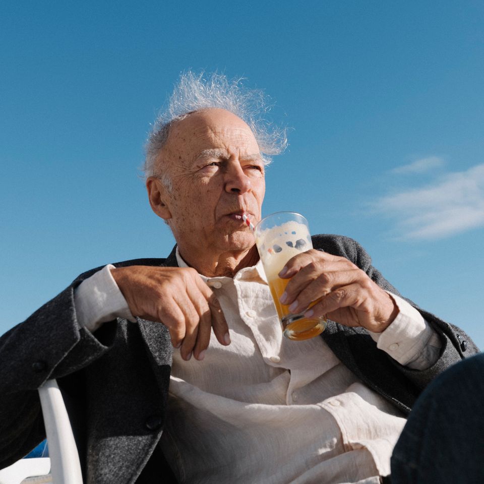 Peter Singer sitzt vor einem strahlend blauen Himmel, trinkt eine orangene Flüssigkeit mit einem Strohhalm aus einem Glas