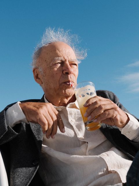 Peter Singer sitzt vor einem strahlend blauen Himmel, trinkt eine orangene Flüssigkeit mit einem Strohhalm aus einem Glas