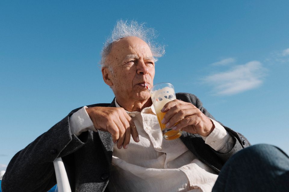 Peter Singer sitzt vor einem strahlend blauen Himmel, trinkt eine orangene Flüssigkeit mit einem Strohhalm aus einem Glas 1.3333