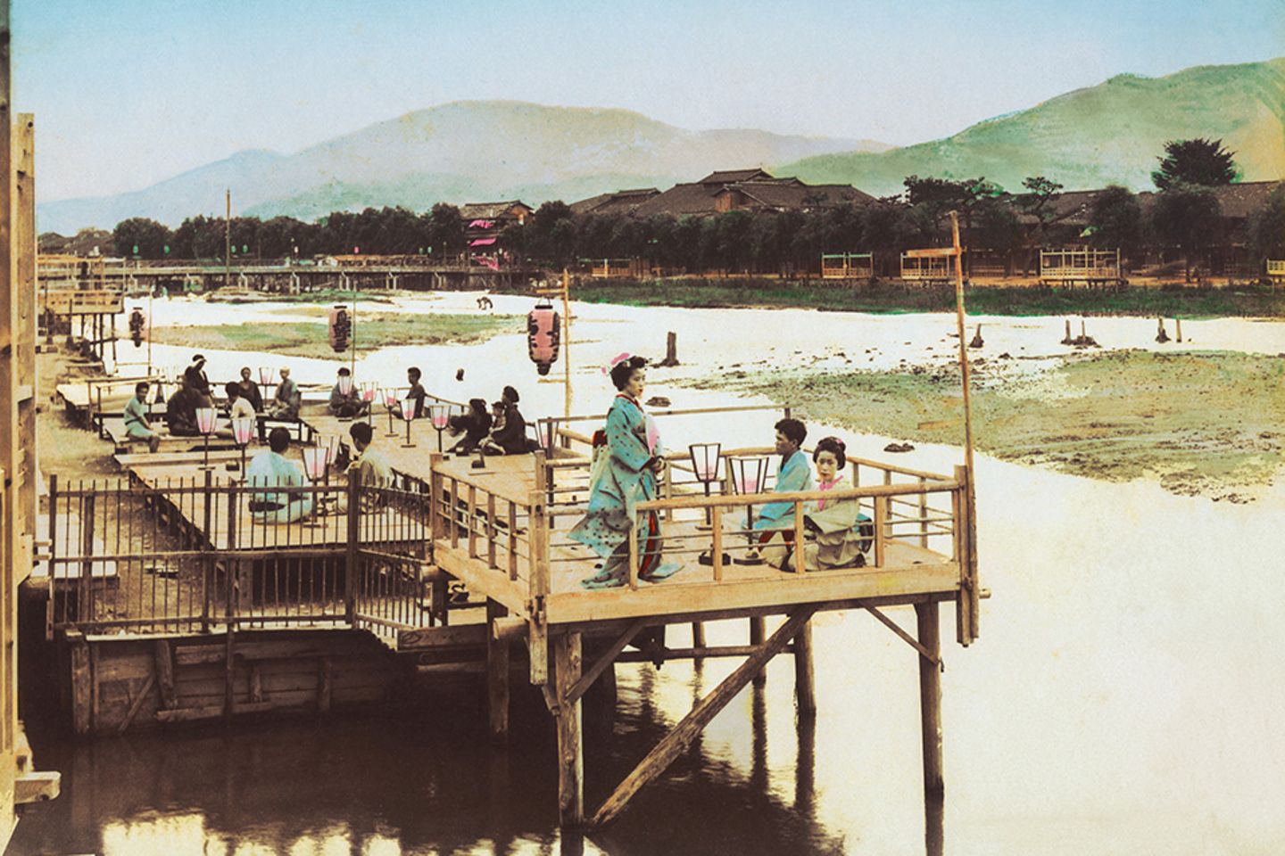 Sommer in Kyoto sind von Hitze und hoher Luftfeuchtigkeit geprägt, kein Wunder: Die Stadt liegt in einem Talbecken, umschlossen von Bergen. Am Fluss gelegene Orte wie Shijō oder Arashiyama sind seit jeher beliebte Treffpunkte, um etwas Abkühlung zu finden. Das gemeinsame Genießen der erfrischenden Abendluft am Flussufer ist eine lang gepflegte Tradition. Das Foto zeigt im Vordergrund elegant gekleidete Geishas, die diesem Sommerritual auf einer der Holzplattformen über dem Wasser nachgehen.