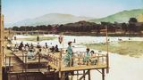 Sommer in Kyoto sind von Hitze und hoher Luftfeuchtigkeit geprägt, kein Wunder: Die Stadt liegt in einem Talbecken, umschlossen von Bergen. Am Fluss gelegene Orte wie Shijō oder Arashiyama sind seit jeher beliebte Treffpunkte, um etwas Abkühlung zu finden. Das gemeinsame Genießen der erfrischenden Abendluft am Flussufer ist eine lang gepflegte Tradition. Das Foto zeigt im Vordergrund elegant gekleidete Geishas, die diesem Sommerritual auf einer der Holzplattformen über dem Wasser nachgehen.