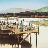 Sommer in Kyoto sind von Hitze und hoher Luftfeuchtigkeit geprägt, kein Wunder: Die Stadt liegt in einem Talbecken, umschlossen von Bergen. Am Fluss gelegene Orte wie Shijō oder Arashiyama sind seit jeher beliebte Treffpunkte, um etwas Abkühlung zu finden. Das gemeinsame Genießen der erfrischenden Abendluft am Flussufer ist eine lang gepflegte Tradition. Das Foto zeigt im Vordergrund elegant gekleidete Geishas, die diesem Sommerritual auf einer der Holzplattformen über dem Wasser nachgehen.