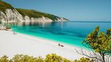 Amazing Fteri beach lagoon, Cephalonia Kefalonia, Greece. Tourists under umbrella relax near clear blue emerald turquise sea water. White rocks in background.