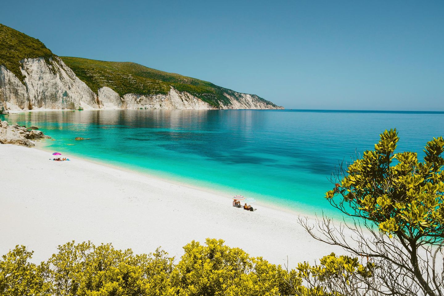 Amazing Fteri beach lagoon, Cephalonia Kefalonia, Greece. Tourists under umbrella relax near clear blue emerald turquise sea water. White rocks in background.
