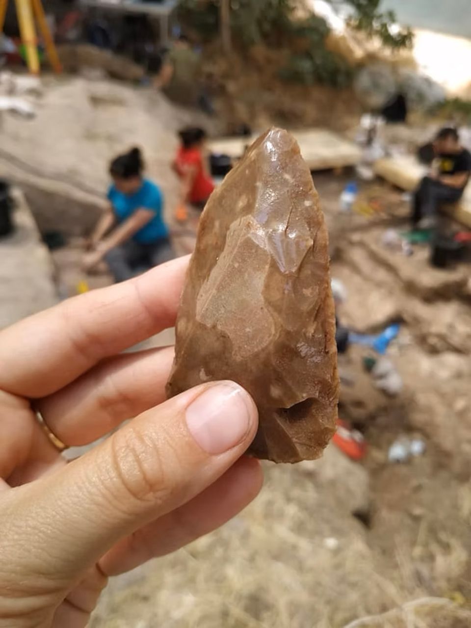 Eine Hand hält ein mehrere Jahrzehntausende altes Steinwerkzeug, dahinter Archäologen bei der Arbeit
