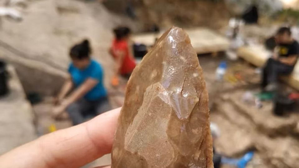 Eine Hand hält ein mehrere Jahrzehntausende altes Steinwerkzeug, dahinter Archäologen bei der Arbeit 