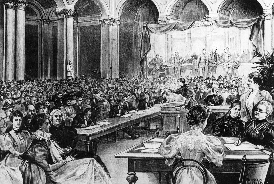 Hunderte Frauen und Männer sitzen in einem prächtigen Saal, an dem eine Rednerin am Pult steht, Illustration, 19. Jh.