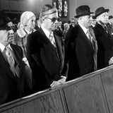 Vier Männer in dunklen Anzügen stehen in einer Bankreihe