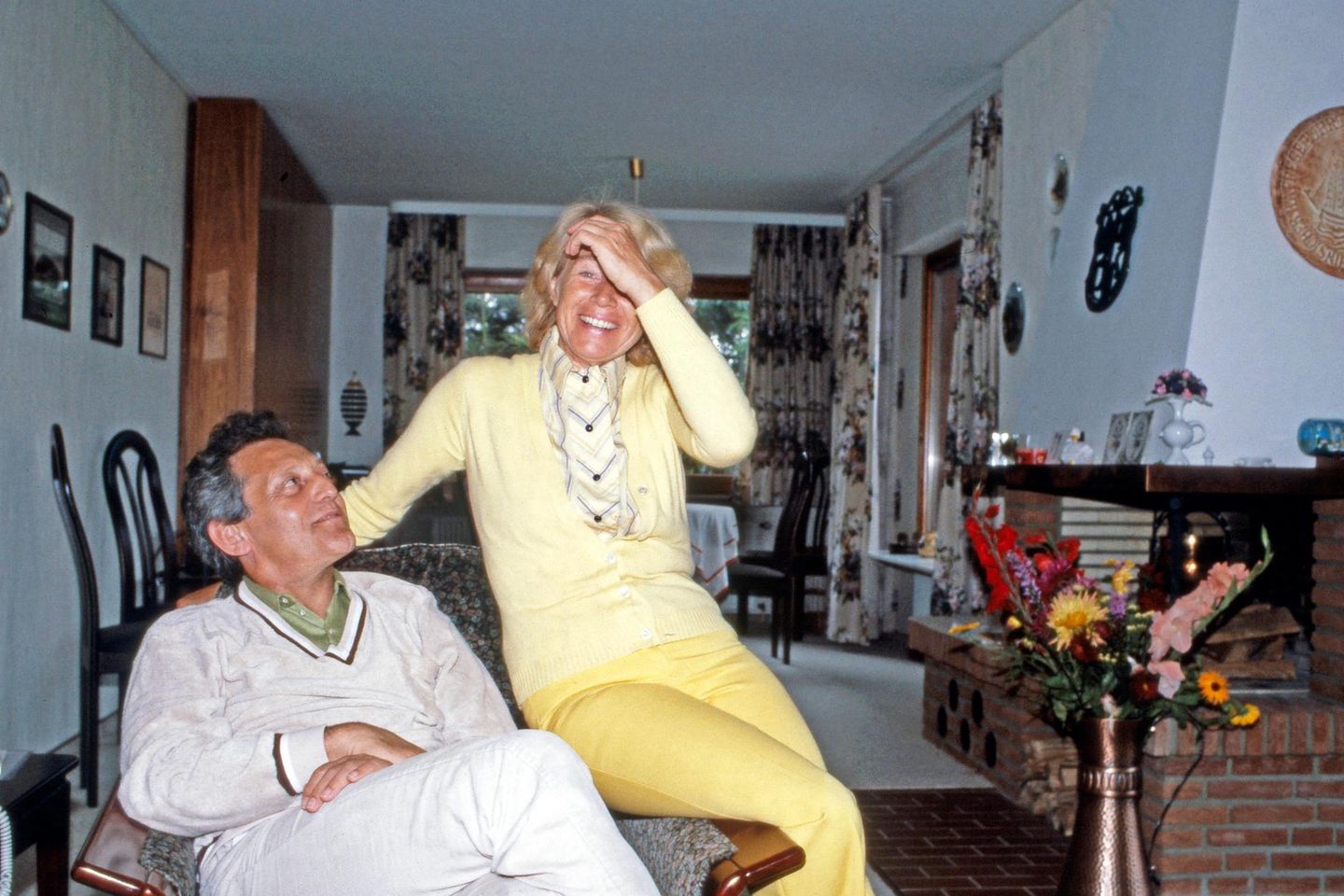 Mann in einem Wohnzimmersessel, auf dessen Lehne sitzt eine lachende Frau