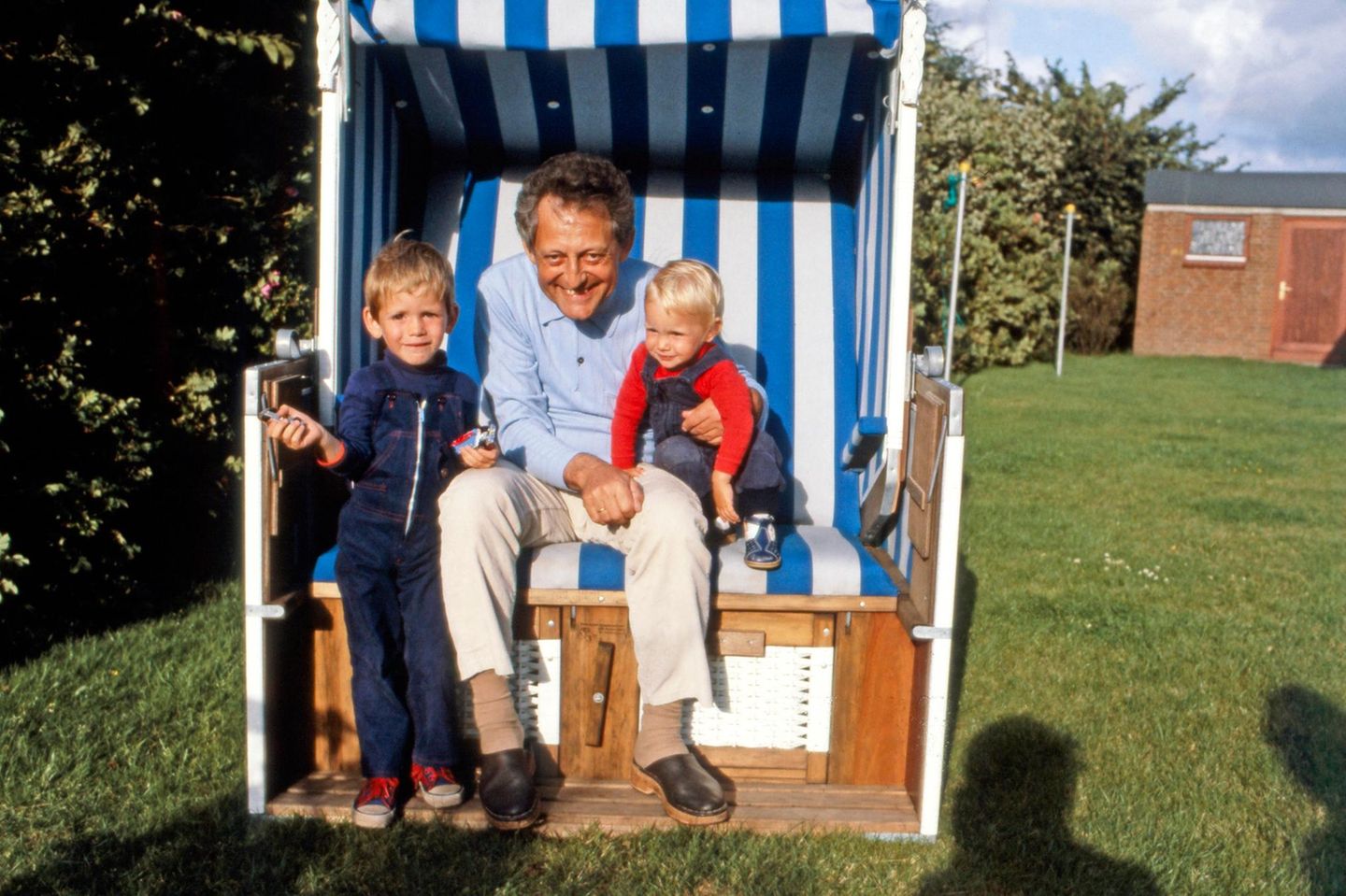 Mann in einem im einem Garten aufgestellten Strandkorb, auf seinem Schoß sitzen zwei kleine Kinder