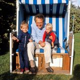 Mann in einem im einem Garten aufgestellten Strandkorb, auf seinem Schoß sitzen zwei kleine Kinder