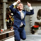 In einen blauen Anzug gekleideter Mann im Luftsprung, die beiden Zeigefinger in die Luft gestreckt