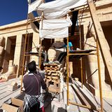 Männer auf einem Baugerüst, darunter ein Statuenkopf auf einem Stapel Vierkanthölzer