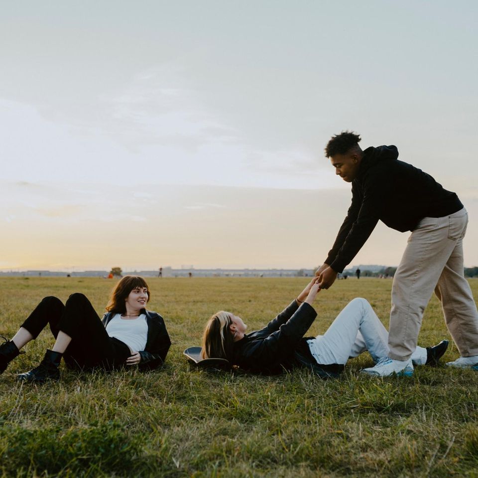 Drei Jugendliche auf einer Wiese, einer hilft seiner Freundin beim Aufstehen