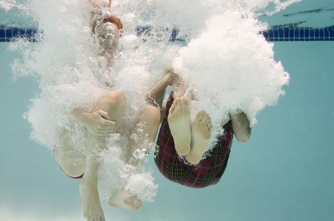 Unterwasseraufnahme: Zwei Menschen mit angwinkelten Beinen kurz nach dem Eintauchen ins Wasser, mit Luftblasen um sie herum.
