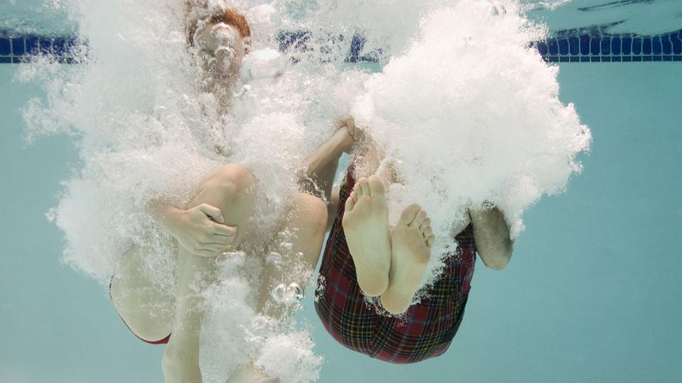 Unterwasseraufnahme: Zwei Menschen mit angwinkelten Beinen kurz nach dem Eintauchen ins Wasser, mit Luftblasen um sie herum.