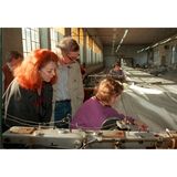 Ein Mann und eine rothaarige Frau sehen einer Näherin bei der Arbeit an Stoffbahnen zu