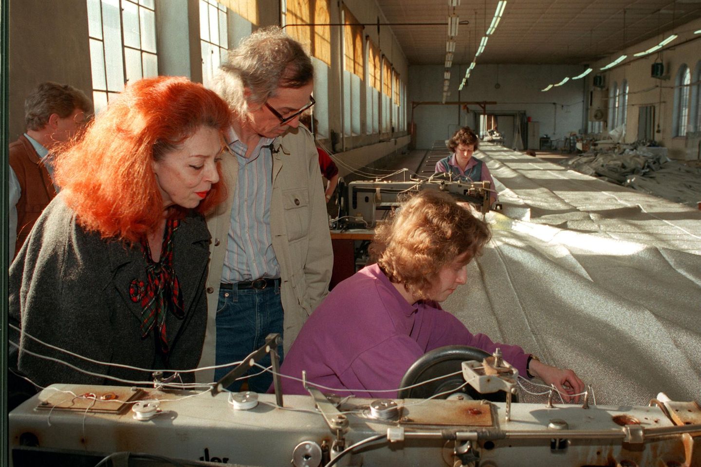 Ein Mann und eine rothaarige Frau sehen einer Näherin bei der Arbeit an Stoffbahnen zu