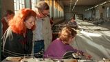 Ein Mann und eine rothaarige Frau sehen einer Näherin bei der Arbeit an Stoffbahnen zu