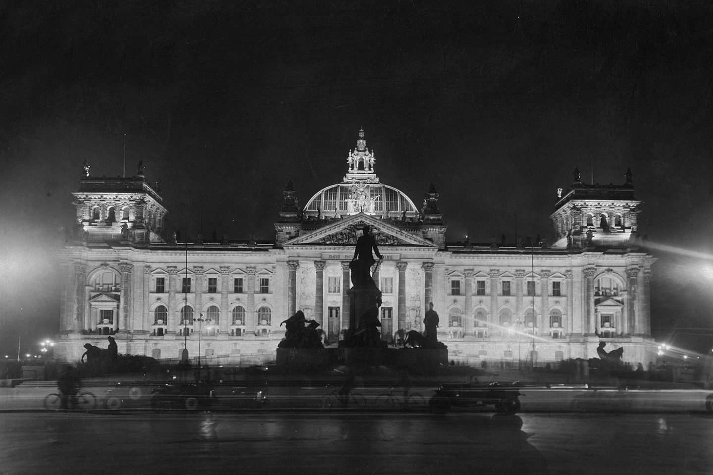 Schwarz-weiß-Aufnahme des erleuchteten Reichstagsgebäudes aus dem Jahr 1928 mit Denkmal davor und Kuppel