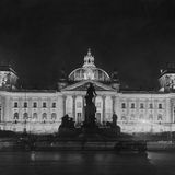 Schwarz-weiß-Aufnahme des erleuchteten Reichstagsgebäudes aus dem Jahr 1928 mit Denkmal davor und Kuppel