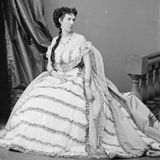 Ganzkörperporträtfoto einer sitzenden Frau in hellem Rüschengewand, mit Schärpe und kunstvoller Frisur, um 1860