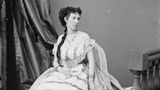 Ganzkörperporträtfoto einer sitzenden Frau in hellem Rüschengewand, mit Schärpe und kunstvoller Frisur, um 1860